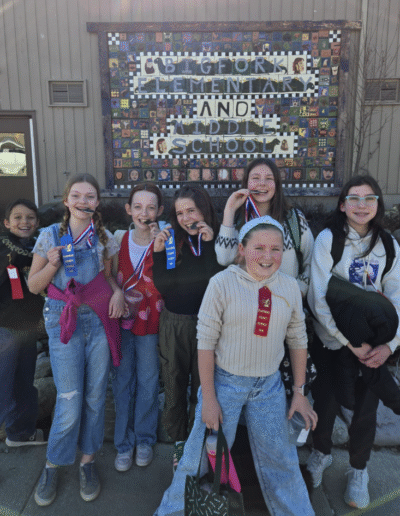A group of fifth graders stand in front of a Bigfork School sign, smiling and holding up their science fair medals and ribbons.